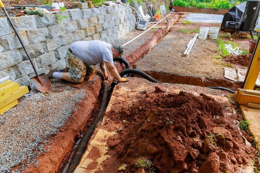 Worker laying corrugated drainage pipe in a trench beside a block retaining wall